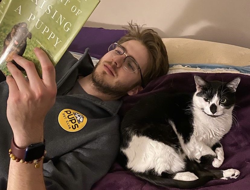 Man reading a book about raising a puppy with a cat lying next to him on a couch.
